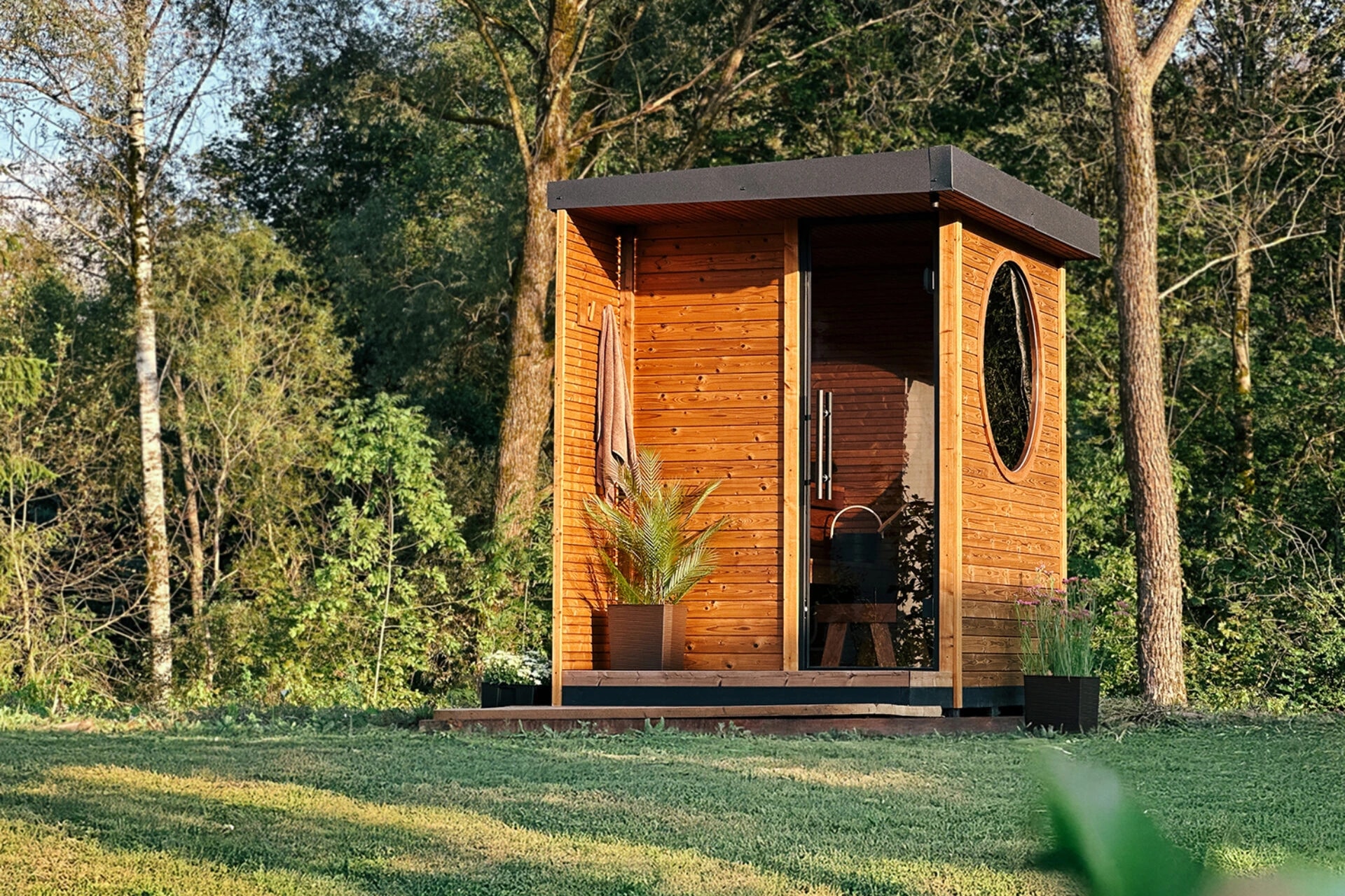 Kaia timber outdoor sauna with a front terrace and circular side window in a wooded garden